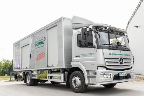 Silberner Transporter der Firma Dommel mit geöffneter Ladefläche auf einem gepflasterten Parkplatz.