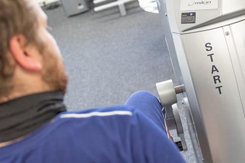 Mann sitzt an einem Fitnessgerät mit Anzeige und drückt auf die Starttaste, im Hintergrund weitere Trainingsgeräte.