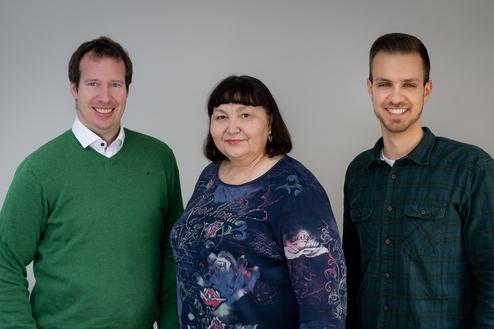 Drei Personen stehen vor einem grauen Hintergrund: zwei Männer in Pullovern und eine Frau in bunter Bluse.