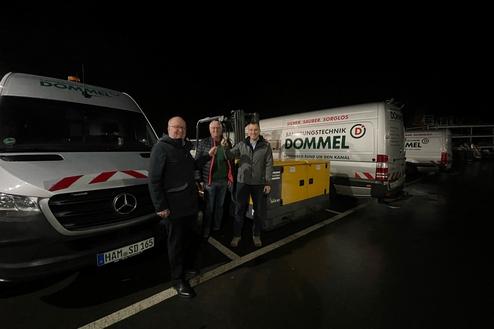 Drei Männer stehen neben einem gelben Generator und zwei Autos der Firma Dommel auf einem dunklen Parkplatz.