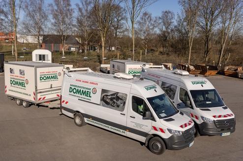 Drei Firmenfahrzeuge von Dommel mit Anhänger stehen auf einem Gelände, umgeben von Bäumen und Bauabbrüchen im Hintergrund.