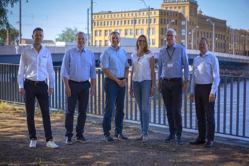 Sechs Personen stehen vor einer Brücke an einem Gewässer, umgeben von urbaner Architektur und Bäume.