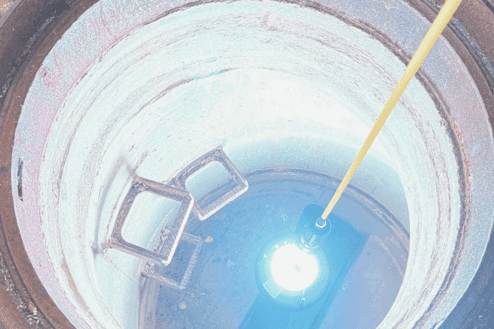 Blick in einen runden Schacht mit einer Lichtquelle und zwei rechteckigen Metallrahmen am Boden.