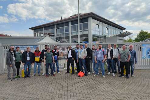 Gruppe von 15 Personen vor einem Gebäude, umgeben von einem Zaun, bei klarer Himmel mit Wolken.