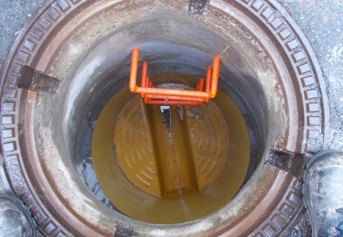 Blick in einen Schacht mit metallischem Boden und orangefarbener Leiter, umgeben von einem runden Gitterdeckel.