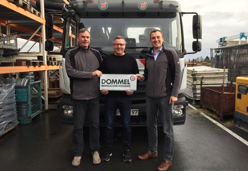 Drei Männer stehen vor einem Lkw und halten ein Schild mit der Aufschrift „DOMMEL NIEDERLASSUNG WIESBADEN“.