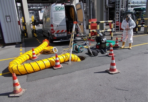 Sanierungsteam in Schutzkleidung arbeitet neben einem offenen Schacht, umgeben von Warnbaken und Arbeitsgeräten.