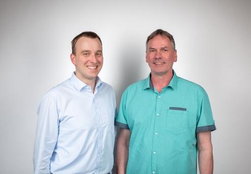 Zwei Männer stehen nebeneinander und lächeln vor einem neutralen Hintergrund. Der Mann links trägt ein hellblaues Hemd, der rechts ein türkisgrünes.