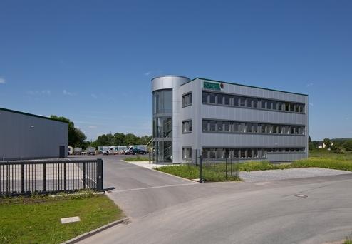 Modernes Bürogebäude mit großen Fenstern und Treppe, umgeben von Wiese und Lagerflächen unter blauem Himmel.
