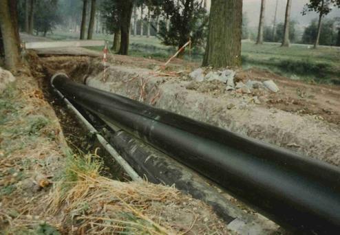 Blick auf eine gegrabene Fläche mit großen, schwarzen Rohren und einer Baustelle, umgeben von Bäumen und Gras.