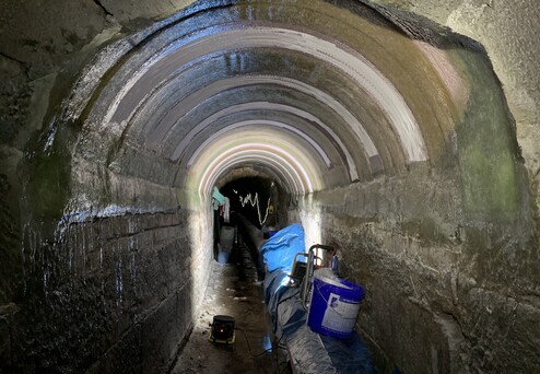 Dunkler Tunnel mit gewölbter Decke, Wasserspuren an der Wand, sowie blauen Planen und Werkzeug am Boden.