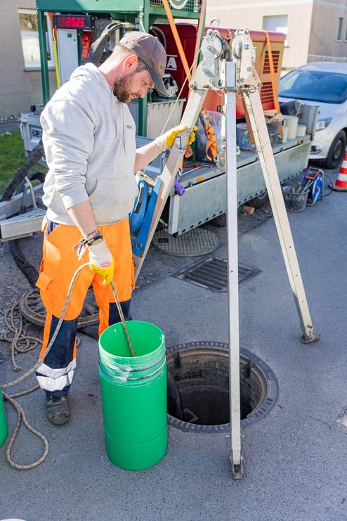 Arbeiter in orangefarbener Arbeitskleidung misst mit einer Rute den Inhalt eines grünen Behälters über einem Schacht.