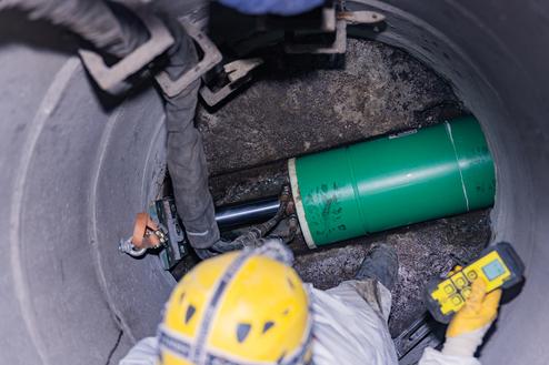 Blick in einen Rohrschacht mit einem grünen Gerät und einem Arbeiter, der ein Handmessgerät hält.