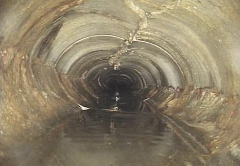 Blick in einen langen, dunklen Abwasserkanal mit Wasseroberfläche und rissigen, unebenen Wänden.