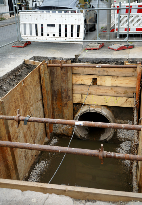 Baugrube mit Holz- und Metallstützen, Wasseransammlung und unterirdischem Rohr im Hintergrund; Baustellensituation.