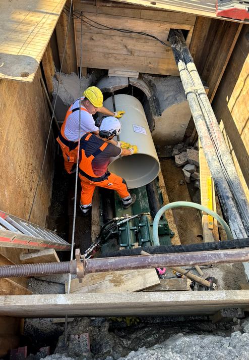 Zwei Arbeiter in Sicherheitskleidung heben ein großes Rohr in einer Baugrube mit schwerem Gerät und Holzverkleidung.