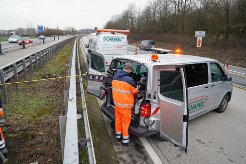 Techniker in orangefarbener Arbeitskleidung arbeitet an einem Firmenfahrzeug an der Autobahn. Warnlichter sind eingeschaltet.