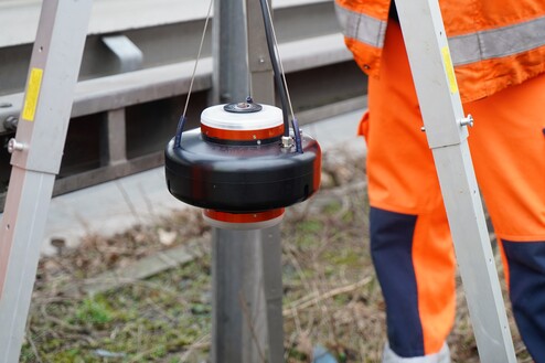 Gerät zur Vermessung hängt an einer Halterung neben einer Aluminiumleiter und einem Arbeiter in orangefarbener Kleidung.