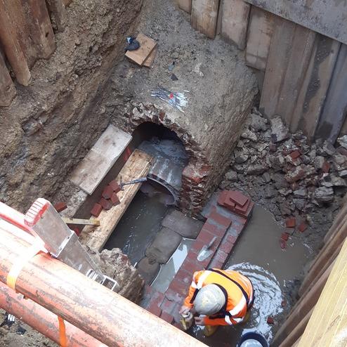 Blick auf eine Baugrube mit einem Arbeiter, Ziegelsteinen und Wasser, umrahmt von Holzverkleidung und Werkzeugen.