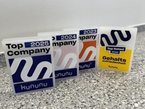 Vier Auszeichnungen von kununu, darunter &bdquo;Top Company&ldquo; und &bdquo;Top Rated&ldquo;, auf einem grauen Boden.