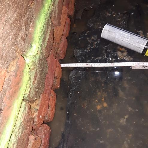 Nahaufnahme einer Ziegelwand mit Riss, einem Messband und Wasserpfütze im Vordergrund.