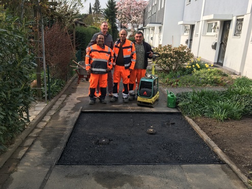 Vier Arbeiter in orangefarbenen Arbeitsanz&uuml;gen stehen vor einer frisch asphaltierten Fl&auml;che in einem Gartenbereich.
