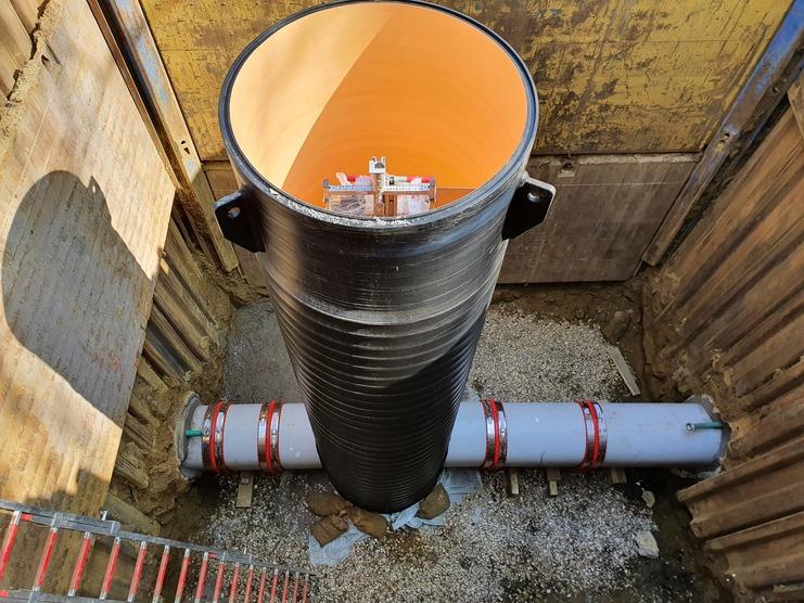 Blick in einen tiefen Schacht mit einem schwarzen Rohr und einer grauen Rohrleitung im Boden, umgeben von Erdmaterial.