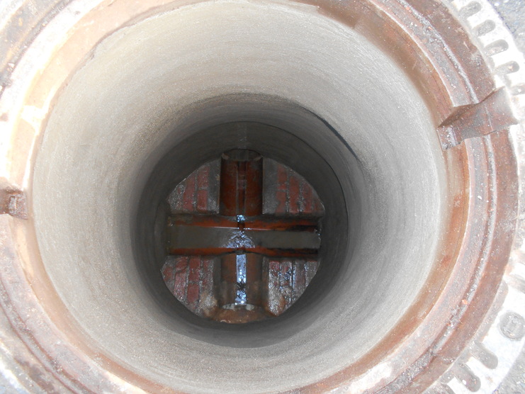 Blick in einen zylindrischen Schacht mit runder Betonwand und querliegendem Metallrost am Boden.