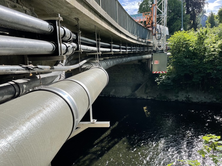 Rohre verlaufen unter einer Brücke über einen Fluss, umgeben von Bäumen und strahlendem Sonnenlicht.