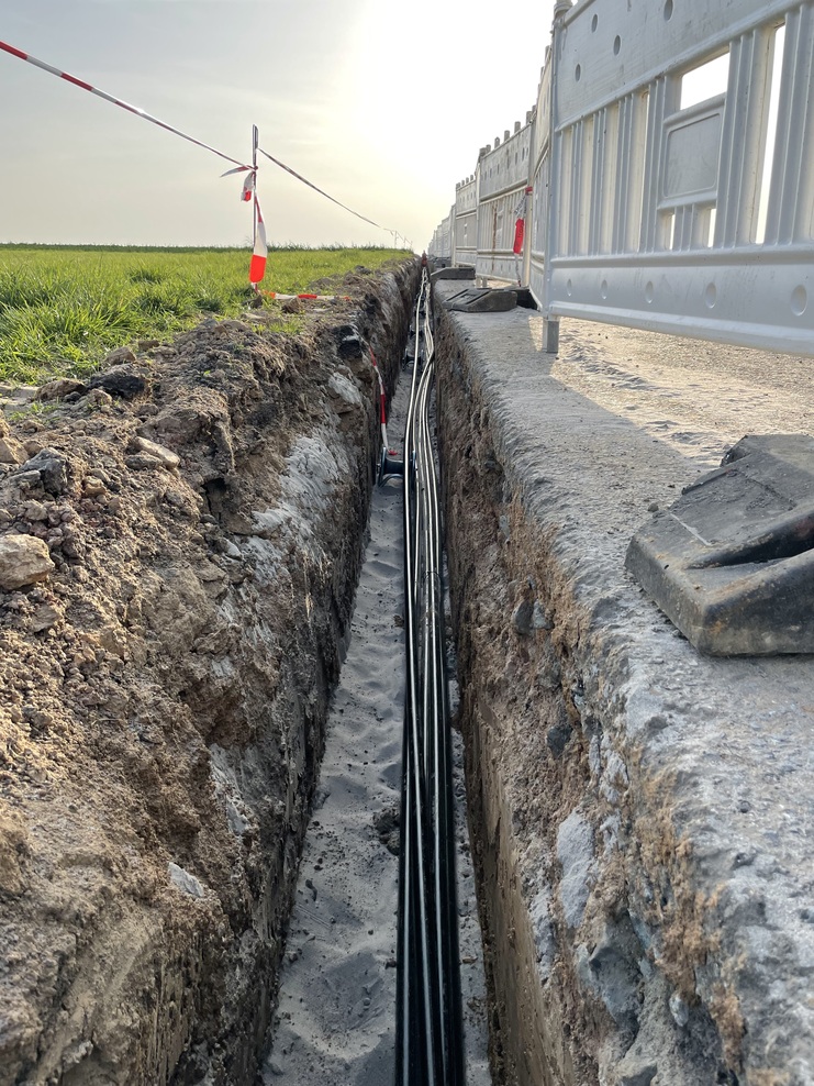 Baugrube mit schwarzen Kabeln, umgeben von Baustellensicherung und grünem Feld im Hintergrund.