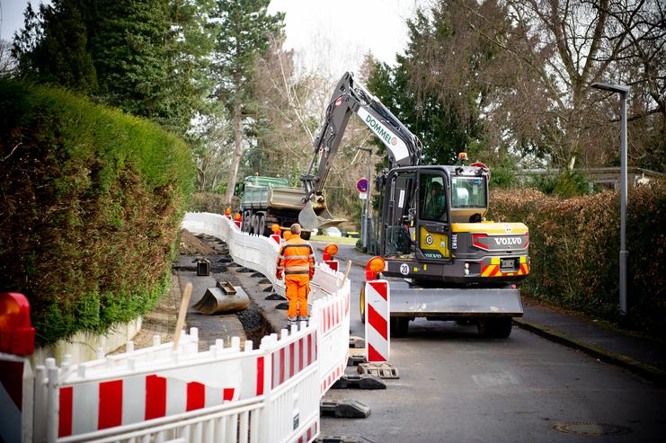 Bauarbeiten an einer Straße mit einem Bagger, Straßenarbeitern und einer Baustellenabsperrung.