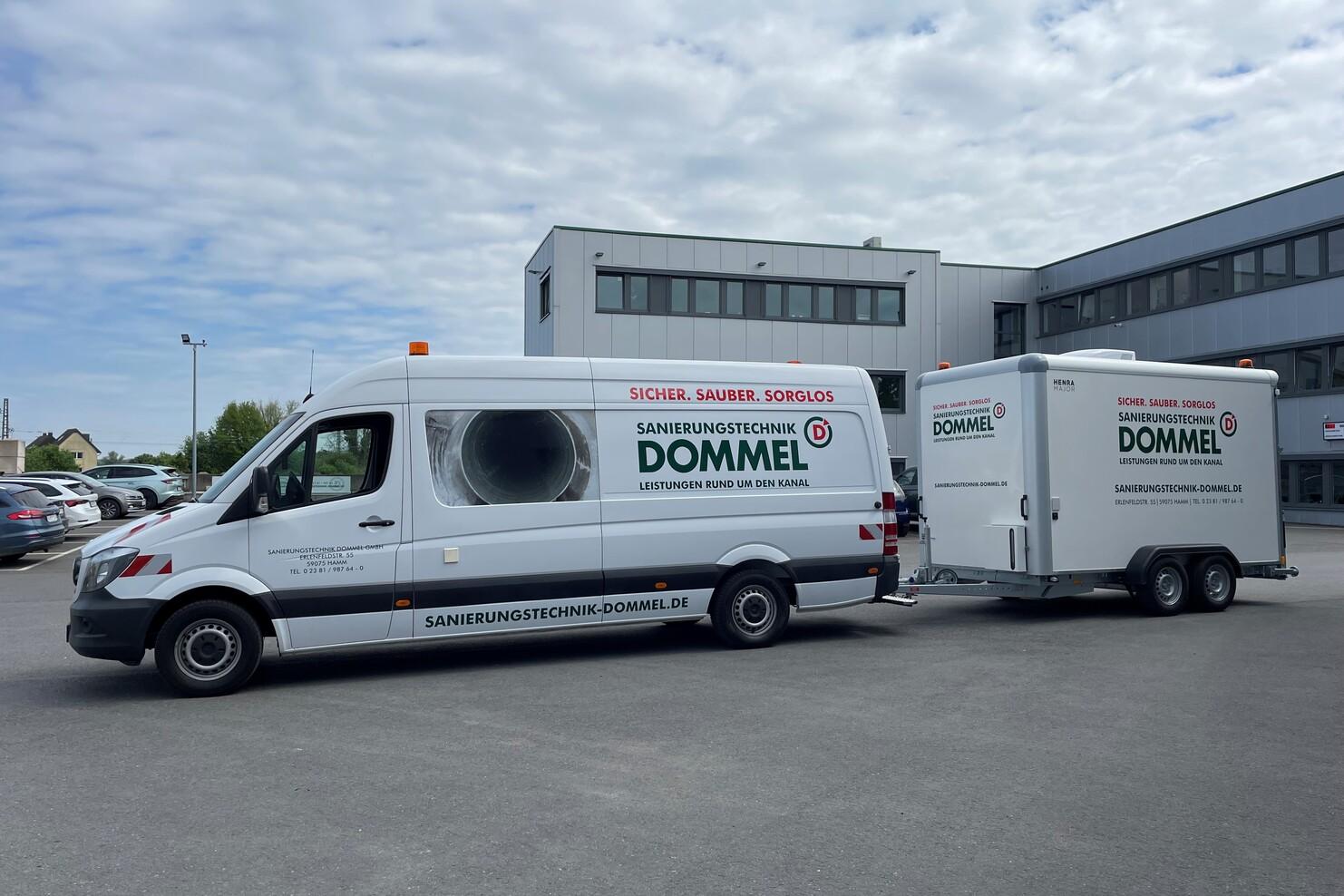 Transporter und Anhänger der Sanierungstechnik Dommel stehen auf einem Parkplatz vor einem Firmengebäude.