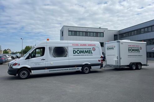 Transporter und Anhänger der Sanierungstechnik Dommel stehen auf einem Parkplatz vor einem Firmengebäude.