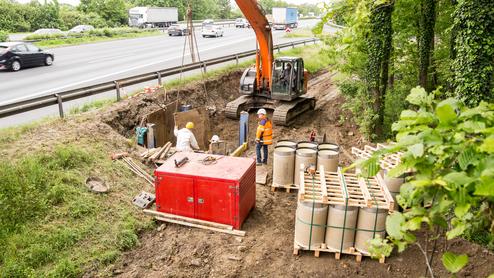 Bauarbeiten an einer Straße mit Bagger und Arbeitern neben einer Baustelle, umgeben von Lkw und Bäumen.
