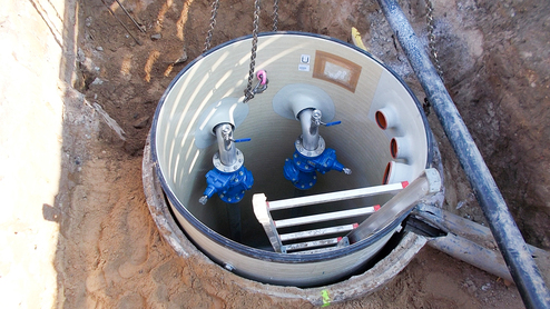 Blick auf einen offenen Schacht mit blauen Ventilen und einer Leiter im inneren. Sandboden um den Schacht herum.