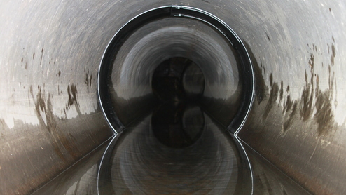 Blick in einen langen, runden Abwasserkanal mit reflektierendem Wasser und glänzenden, metallischen Wänden.