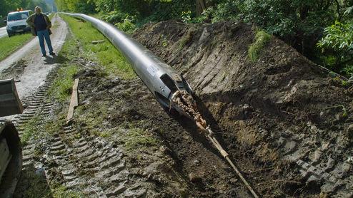 Ein Mann steht neben einer langen Metallröhre, die in einem Graben neben einer Straße liegt.