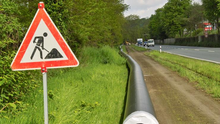 Bauarbeiten-Schild neben einer langen, grauen Rohrleitung an einer grünen Wiese und einer befahrenen Straße.