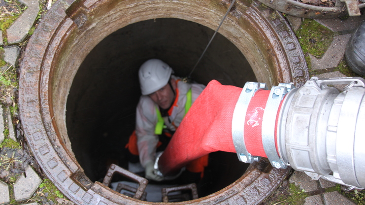 Arbeiter in einem engen Schacht mit einem roten Schlauch, der in den Schacht führt, umgeben von Steinplatten.