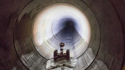 Roboterfahrzeug auf einem Wasserfluss in einem großen zylindrischen Tunnel, Licht am Tunnelende sichtbar.