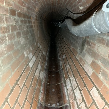 Blick in einen langen, gefliesten Tunnel mit einem grauen Rohr an der Wand und leicht feuchtem Boden.