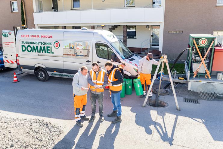 Vier Personen in Warnwesten stehen am Straßenrand und betrachten Pläne, während ein Leiter und technische Ausrüstung bereitstehen.