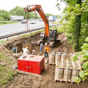 Bauarbeiter neben einer Grube, während ein Bagger Rohre hebt, mit Fahrzeugen auf der Straße im Hintergrund.
