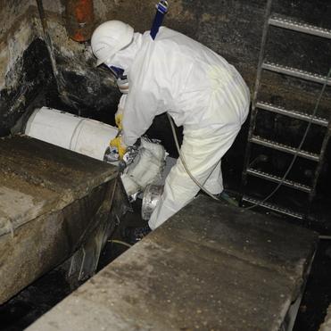 Arbeiter in Schutzanzug repariert eine Rohrleitung in einem stark verschmutzten, engen Kanalraum.