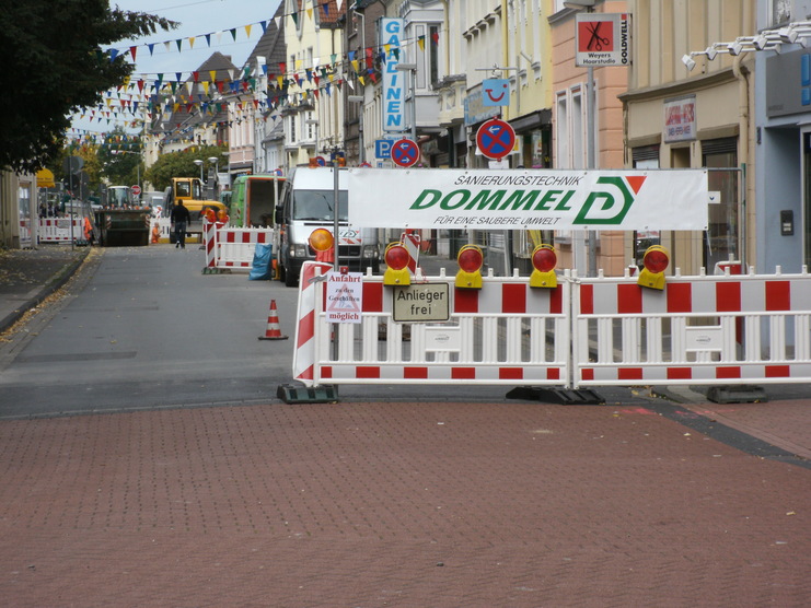 Bauschild und Absperrungen auf einer Baustelle in einer Stadtstraße mit bunten Fahnen im Hintergrund.