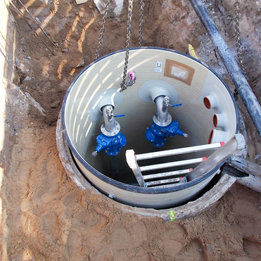 Blick auf einen offenen Schacht mit zwei blauen Ventilen und einer Leiter im Inneren. Sandboden um den Schacht herum.