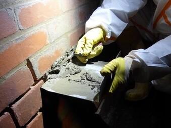 Hände in Schutzanzug mit gelben Handschuhen bearbeiten Mauerwerk mit Mörtel in einer dunklen Umgebung.