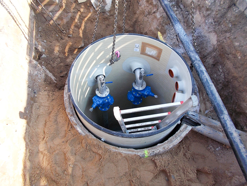 Blick auf einen offenen Schacht mit zwei blauen Ventilen und einer Leiter im Inneren. Sandboden um den Schacht herum.
