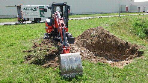 Bagger arbeitet auf einer Wiese, neben einem Lkw mit Aufschrift "Dommel" und einem ausgehobenen Erdloch.