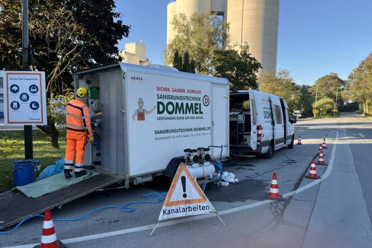 Baufahrzeug mit Sanierungstechnik, Arbeiter in Warnkleidung und Warnschild „Kanalarbeiten“ auf der Straße.